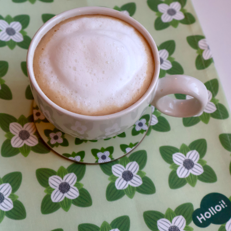 Grønt kjøkkenhåndkle med kaffeblomst-motiv brukt som underlag for en kaffekopp med melkeskum og et matchende glassunderlag