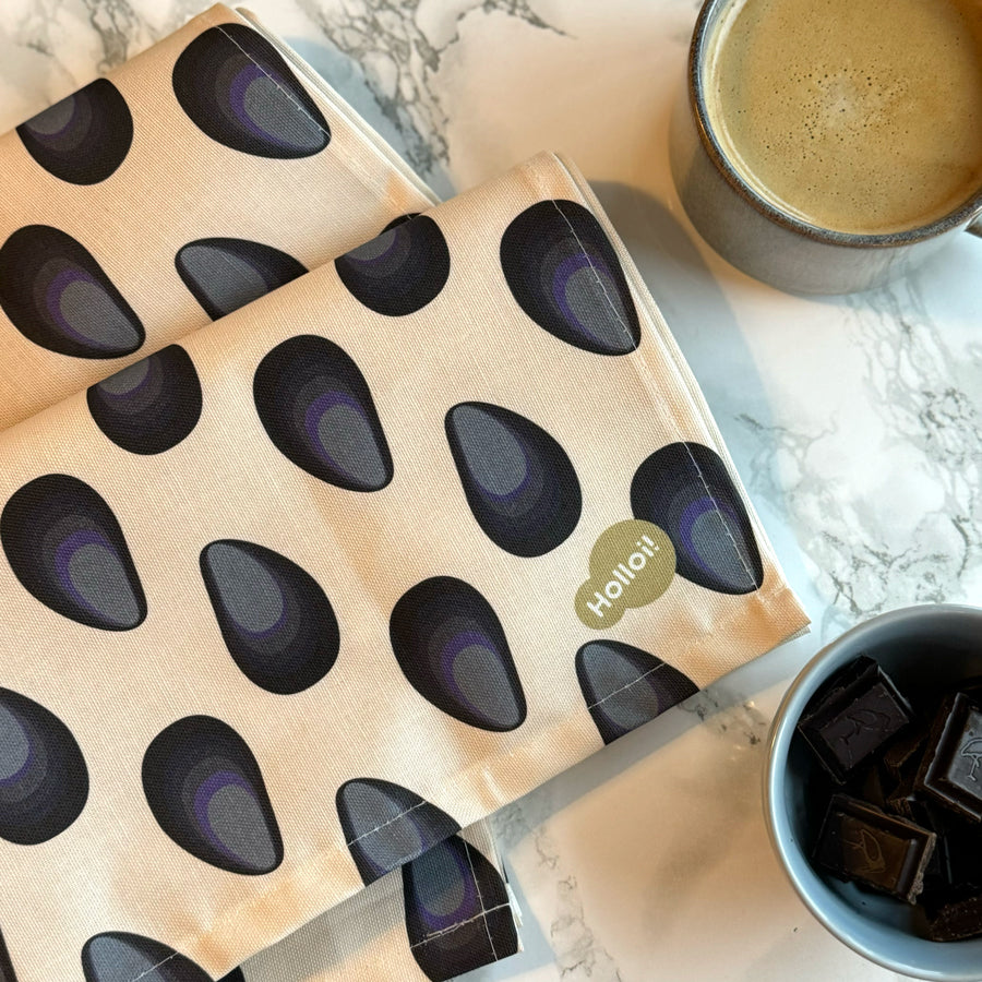 Kjøkkenhåndkle med blåskjell-motiv fra Holloi liggende på kjøkkenbenk sammen med en kaffekopp og en skål sjokolade