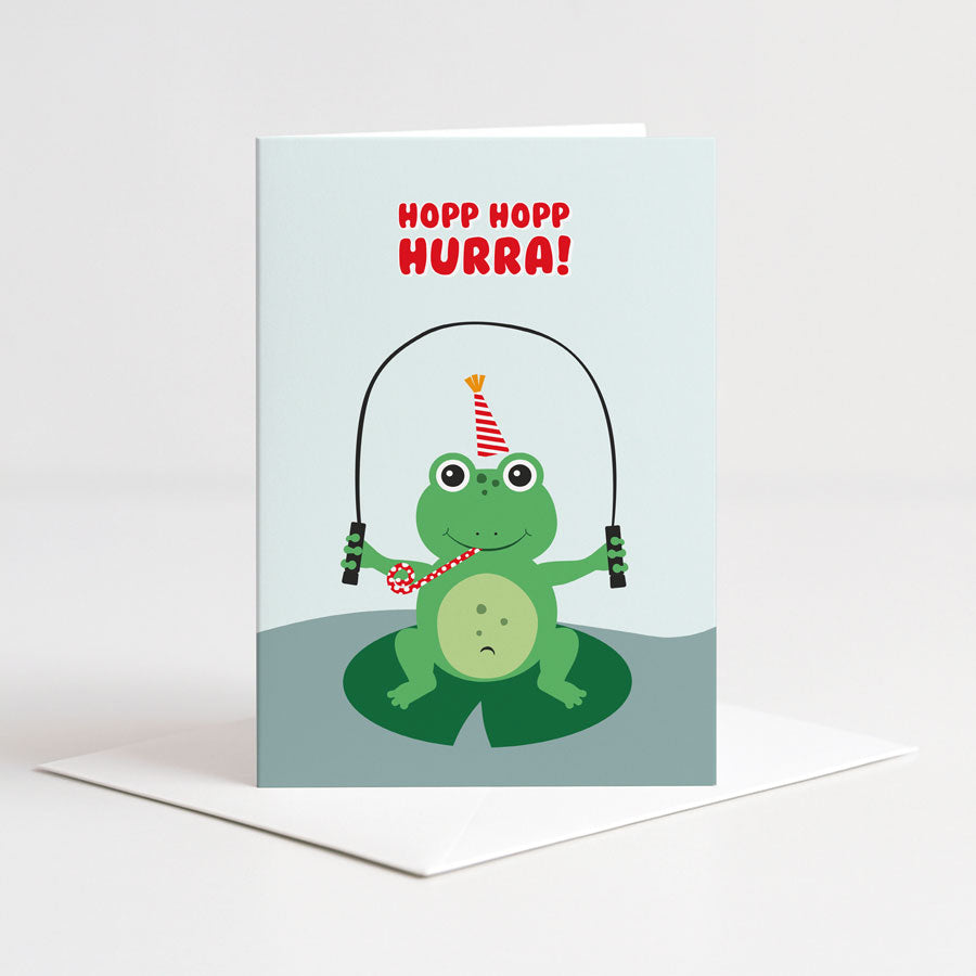Lyseblått dobbelt kort fra Holloi med illustrasjon av en grønn frosk i festhatt som hopper tau på et liljeblad, og teksten HOPP HOPP HURRA