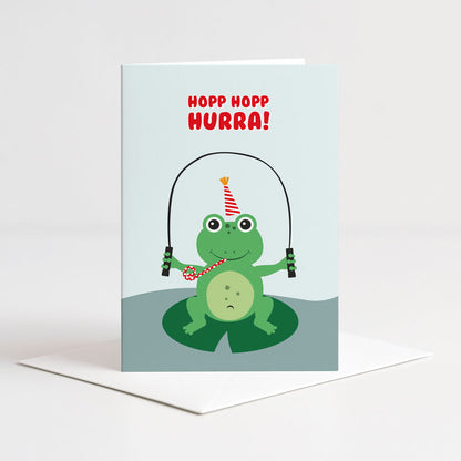 Lyseblått dobbelt kort fra Holloi med illustrasjon av en grønn frosk i festhatt som hopper tau på et liljeblad, og teksten HOPP HOPP HURRA
