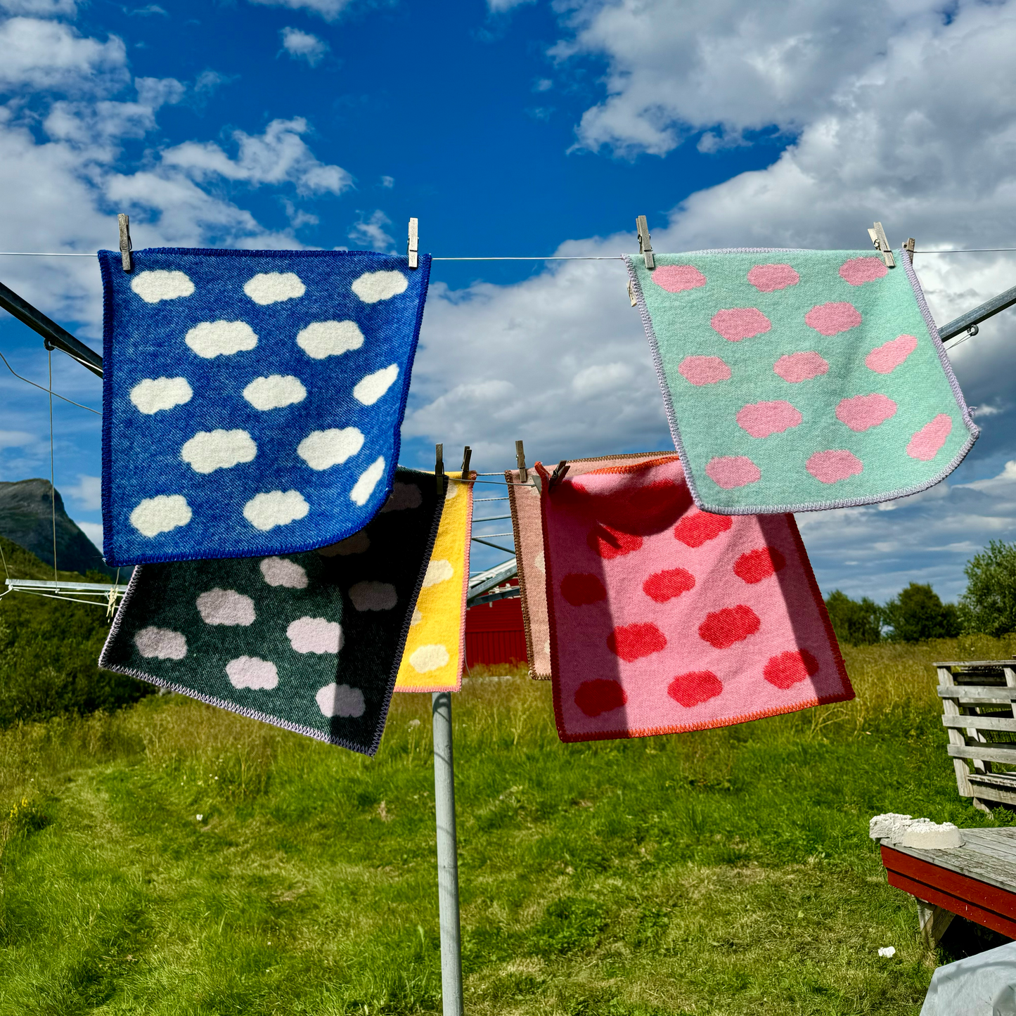 Sitteunderlag i varierte farger, blant annet de blå kalt Godvær, hengende på en snor utendørs i sommerlig vær