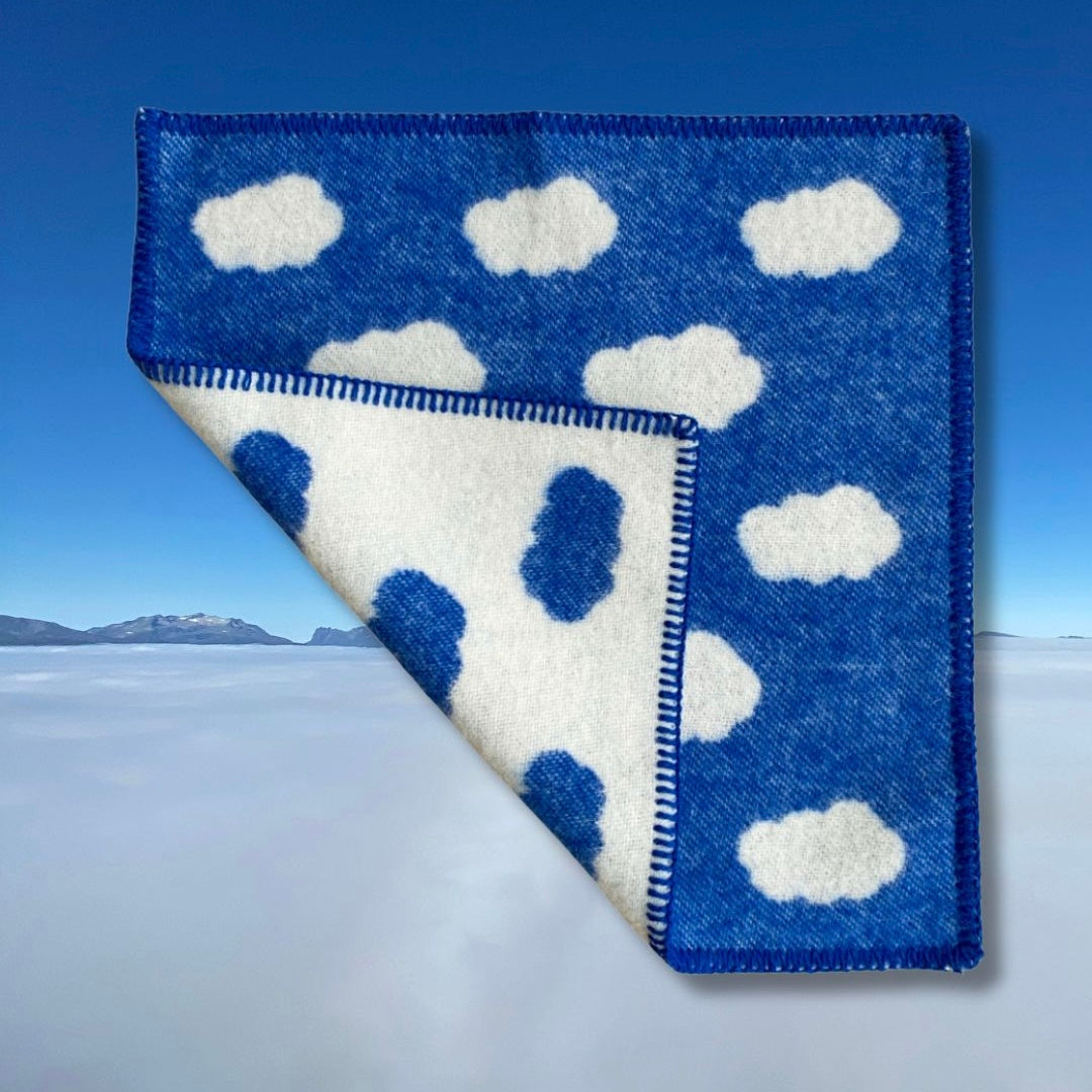 Blått sitteunderlag av ull med et bilde av blå himmel og hvite skyer i bakgrunnen, med en brett som viser forsiden og baksiden på sitteunderlaget