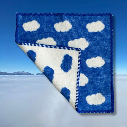 Blått sitteunderlag av ull med et bilde av blå himmel og hvite skyer i bakgrunnen, med en brett som viser forsiden og baksiden på sitteunderlaget