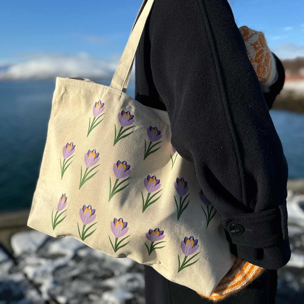 Kvinne som bærer en naturfarget tøyveske med krokusmotiv i fjæra, med stein og hav i bakgrunnen