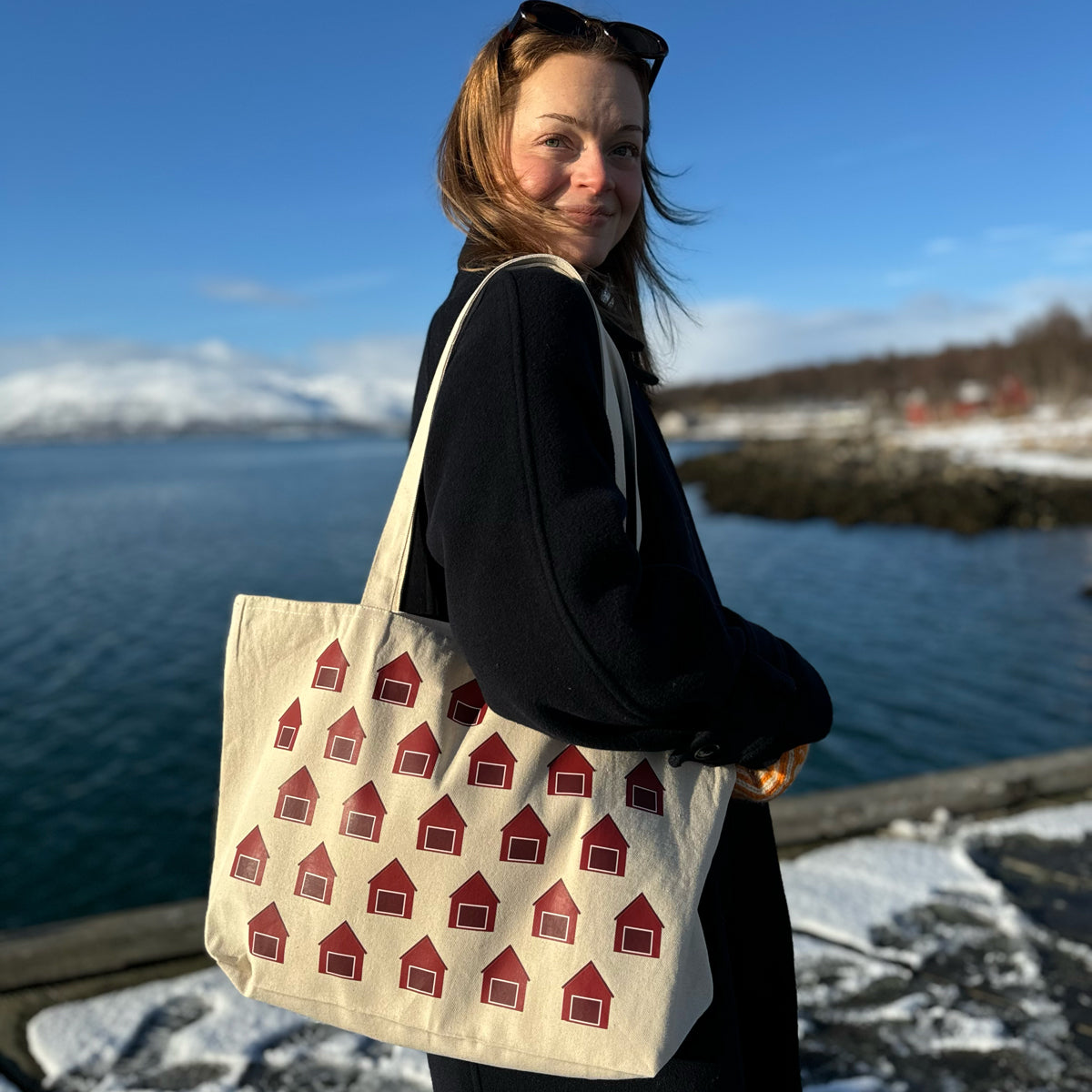 Kvinne med naturfarget tøyveske med naust-motiv over skulderen, fotografert utendørs i kystlandskap
