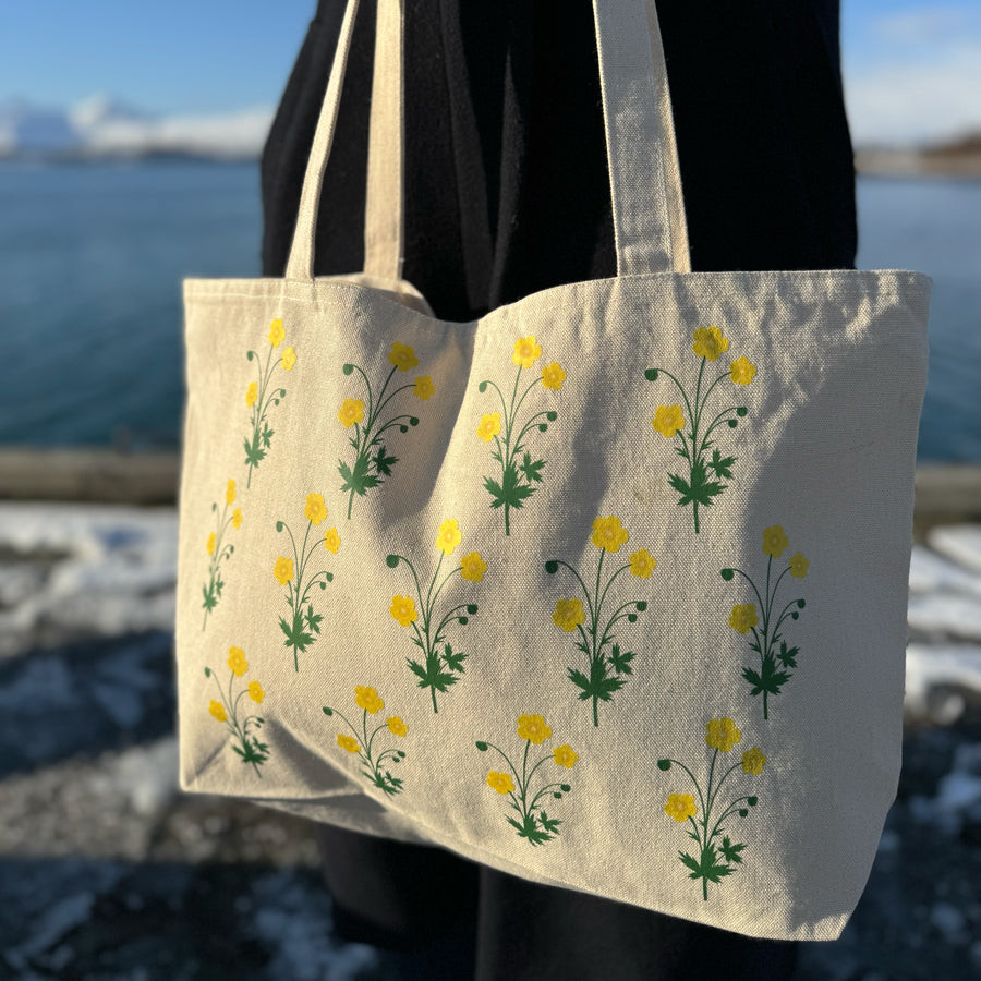 Tøyveske med gult smørblomst-motiv fotografert utendørs mot en bakgrunn av blått hav og kystlandskap