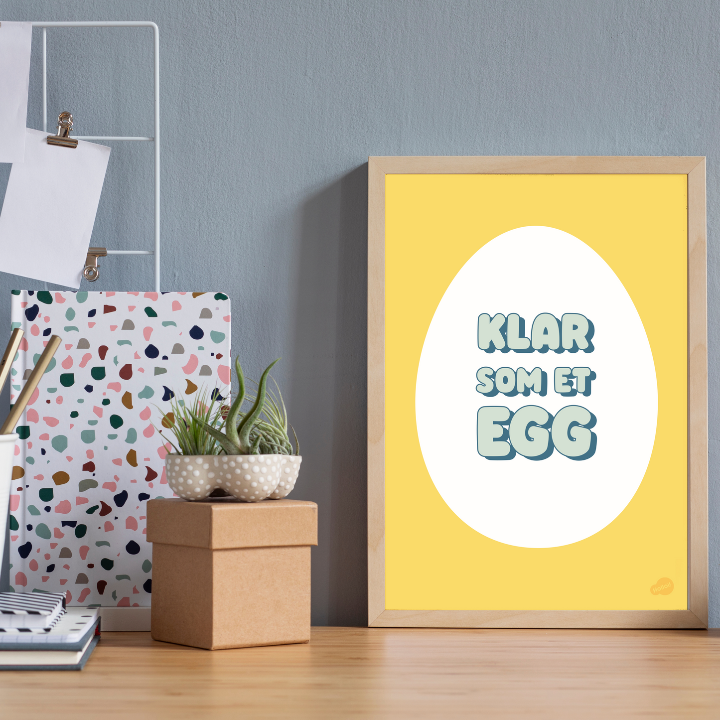 Plakat med illustrasjon av et egg og teksten "Klar som et egg" stående innrammet på en skrivepult